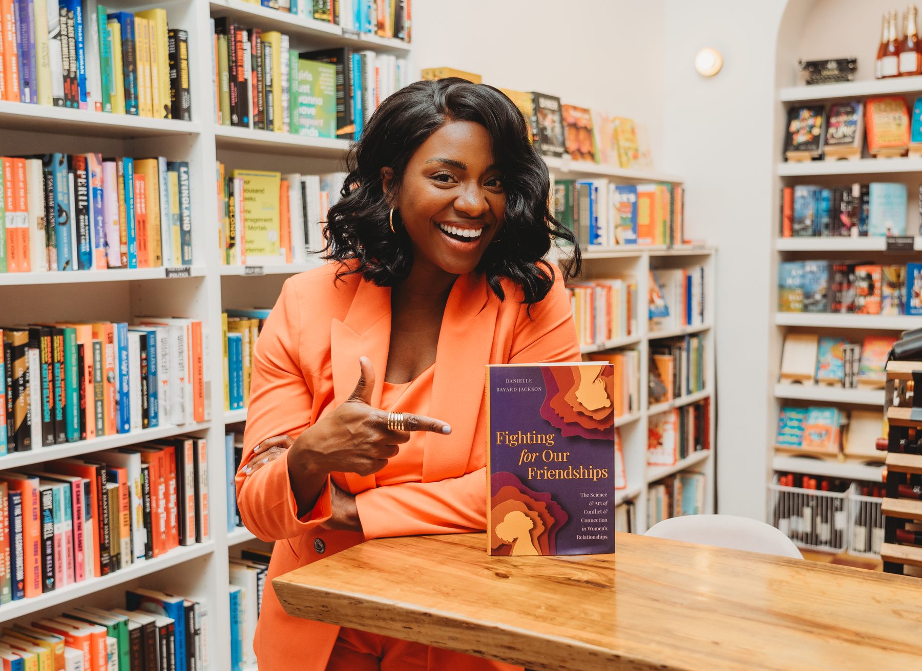 Danielle in an orange suit at a bookstore table, pointing at her book