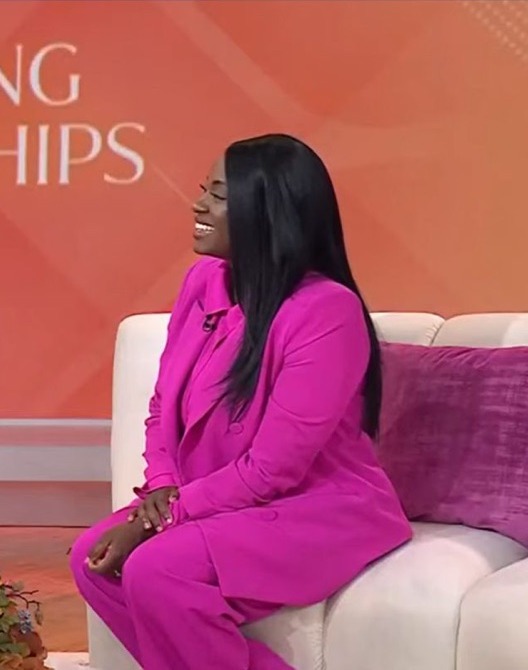 Danielle in a pink suit, seated on a studio couch